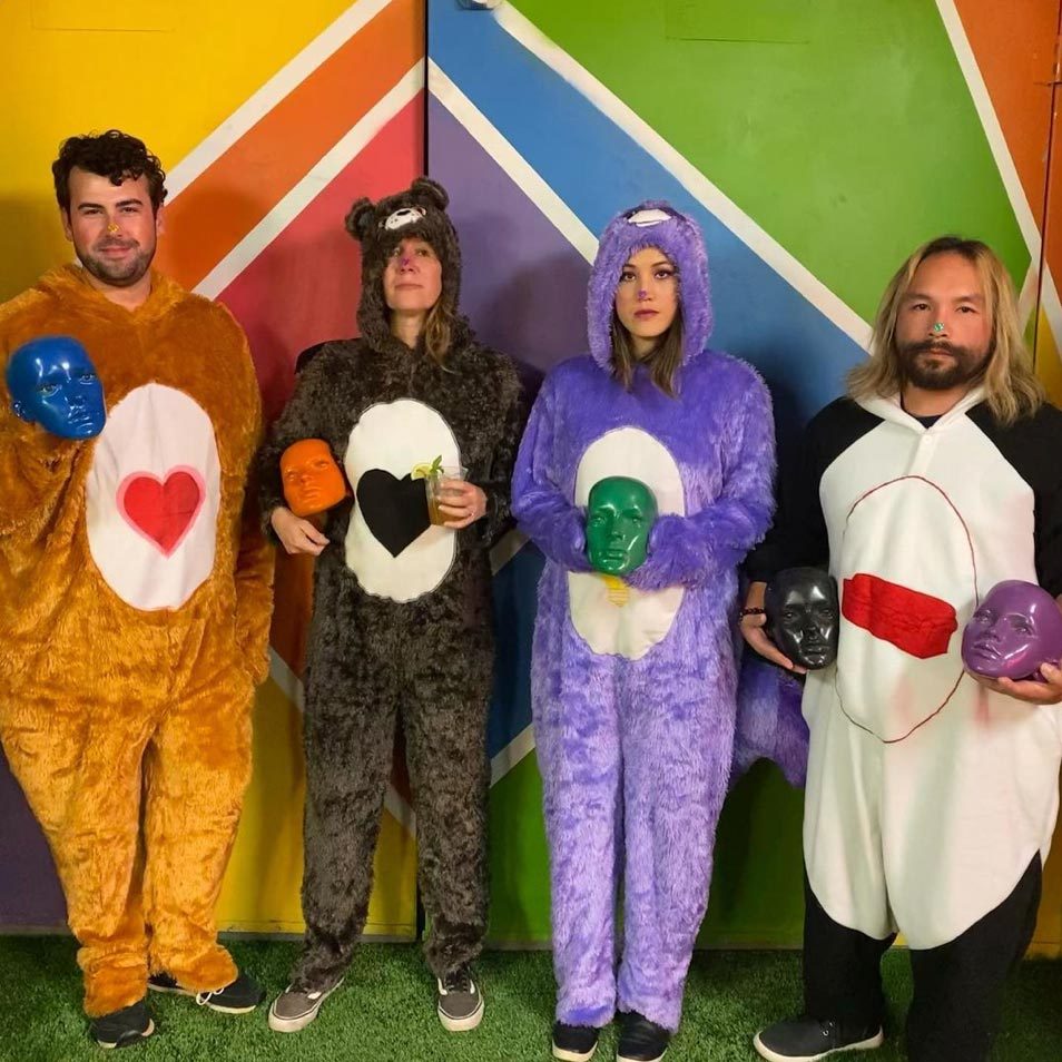Four people wearing colorful bear costumes hold painted masks against a vibrant geometric background.