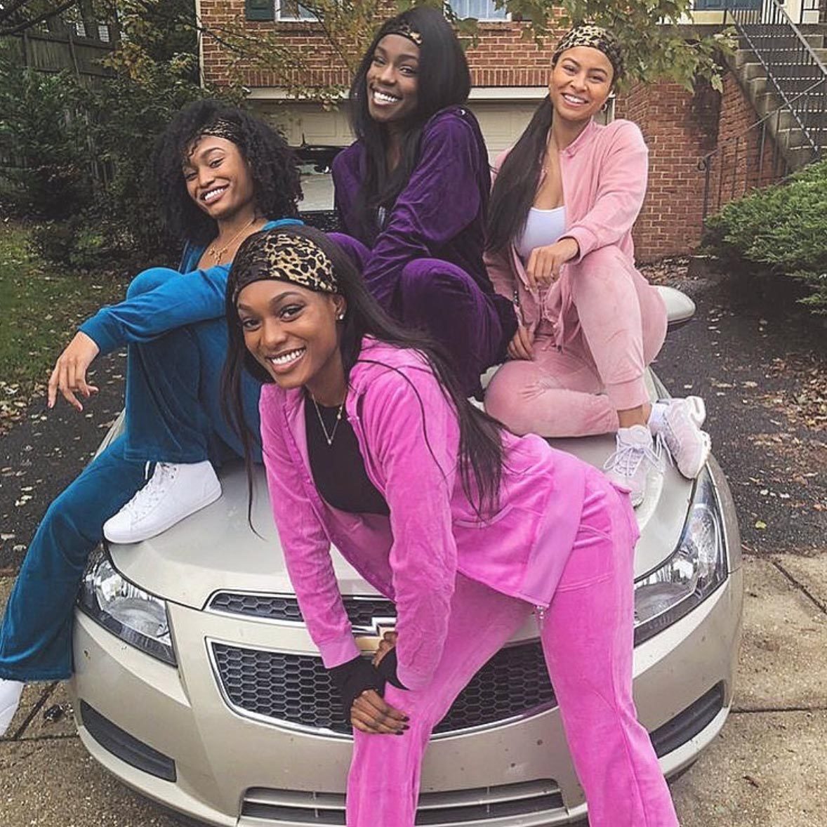 Four people sit and smile on a car's hood, wearing colorful tracksuits, in a suburban driveway surrounded by greenery and a brick house.