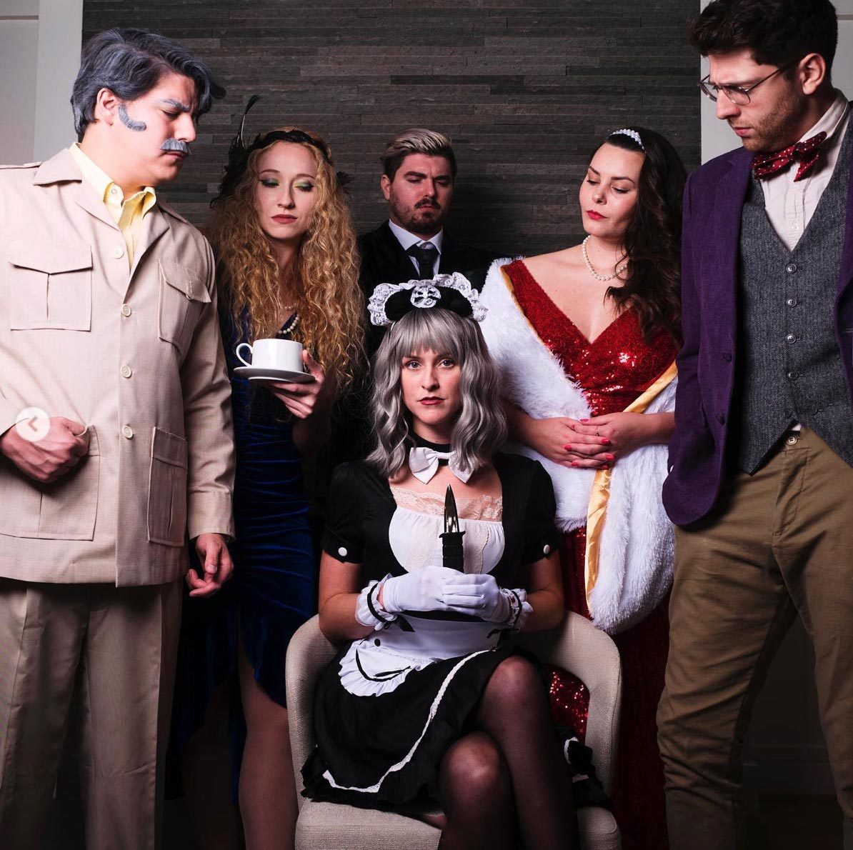 Seated maid holds dagger amidst five people in costume. They're dressed in vintage or formal attire, standing closely in an indoor setting.