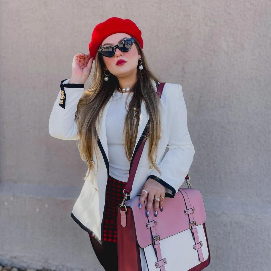 Woman poses stylishly, wearing a red beret and sunglasses, holding a pink bag, standing against a plain, textured wall.