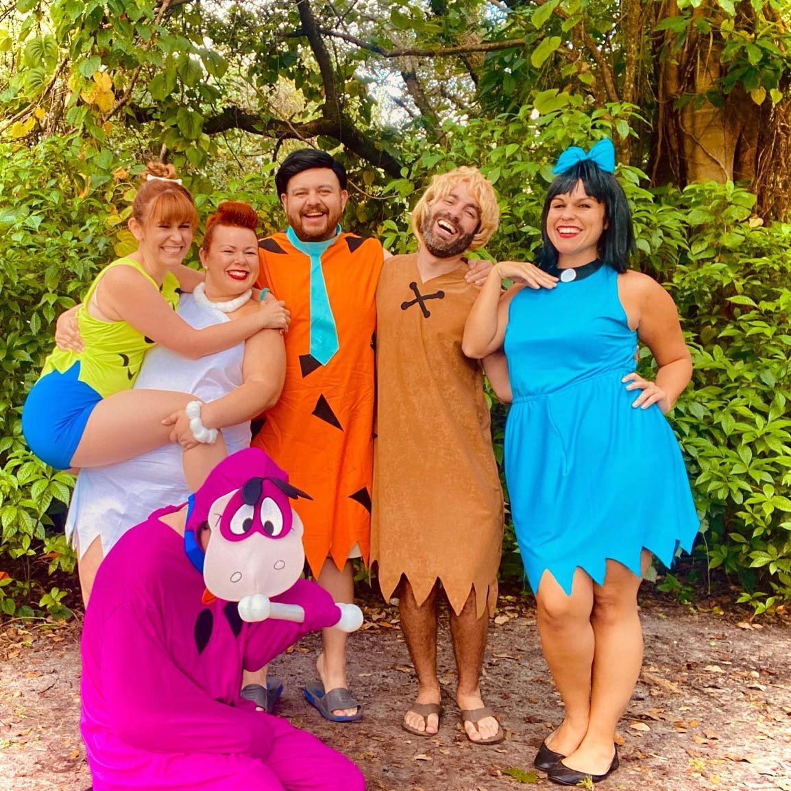 People posing in colorful, Flintstones-themed costumes, smiling under trees in a lush, green outdoor setting.