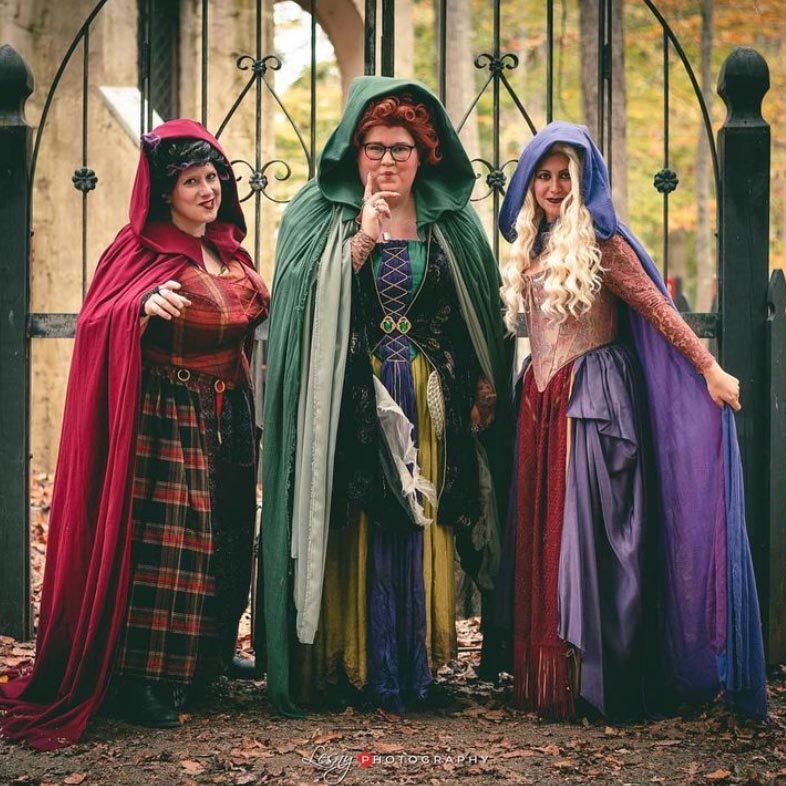 Three people in vibrant medieval costumes stand in front of an iron gate, surrounded by autumn leaves.