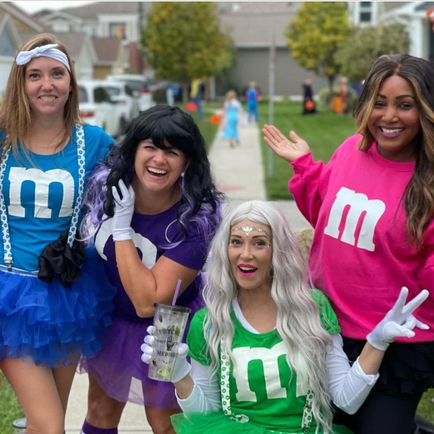 Four women dressed as M&M characters pose, smiling on a suburban sidewalk with houses and costumed people in the background.