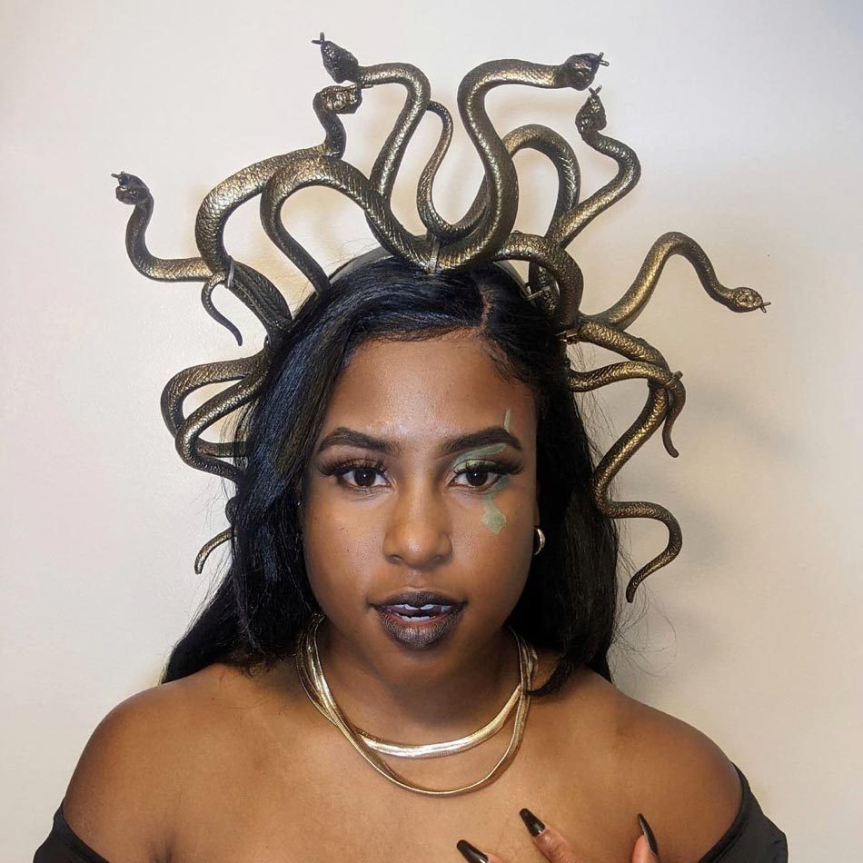 A woman wears a snakes-headpiece and dark makeup, posing against a plain background.