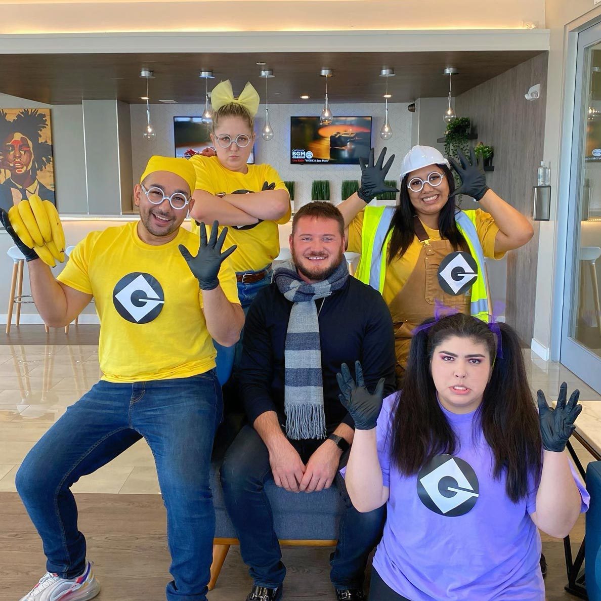 Five people pose in colorful costumes, wearing black gloves, in a modern office lobby featuring artwork and hanging lights.