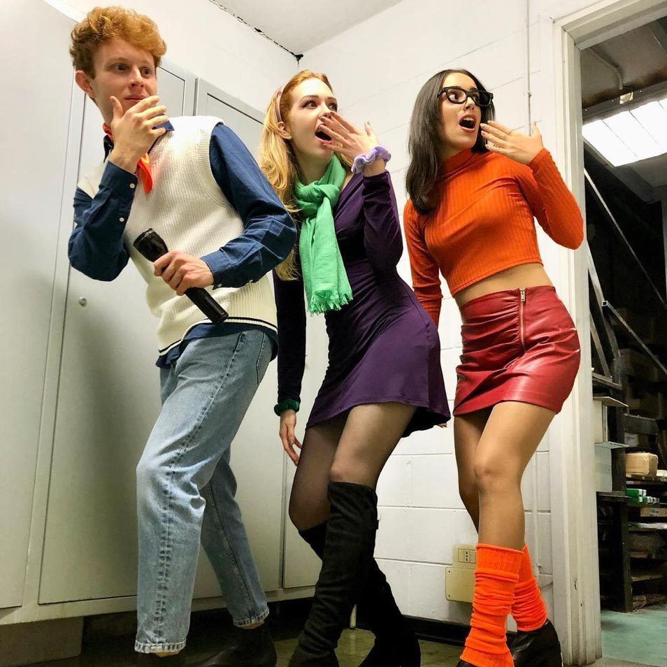 Three people pose dramatically, holding fake surprised expressions, dressed in vibrant, retro-inspired costumes, in a room with white walls and metal cabinets.