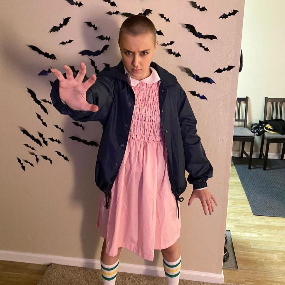 Person extends hand forward, standing in front of wall decorated with black paper bats, wearing pink dress and navy jacket, indoors.