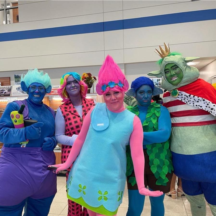 Costumed individuals pose cheerfully in colorful troll-themed outfits, indoors, suggesting a playful event or gathering.
