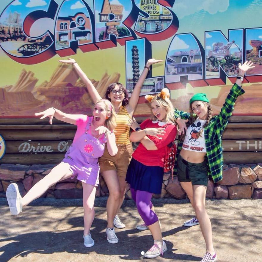 Four people pose energetically in front of a colorful "Cars Land" sign, outdoors, each expressing playful excitement.