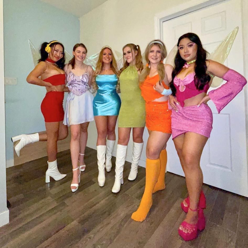 Six women pose in colorful, themed outfits, resembling fairies, inside a room with wooden floors and a closed white door.