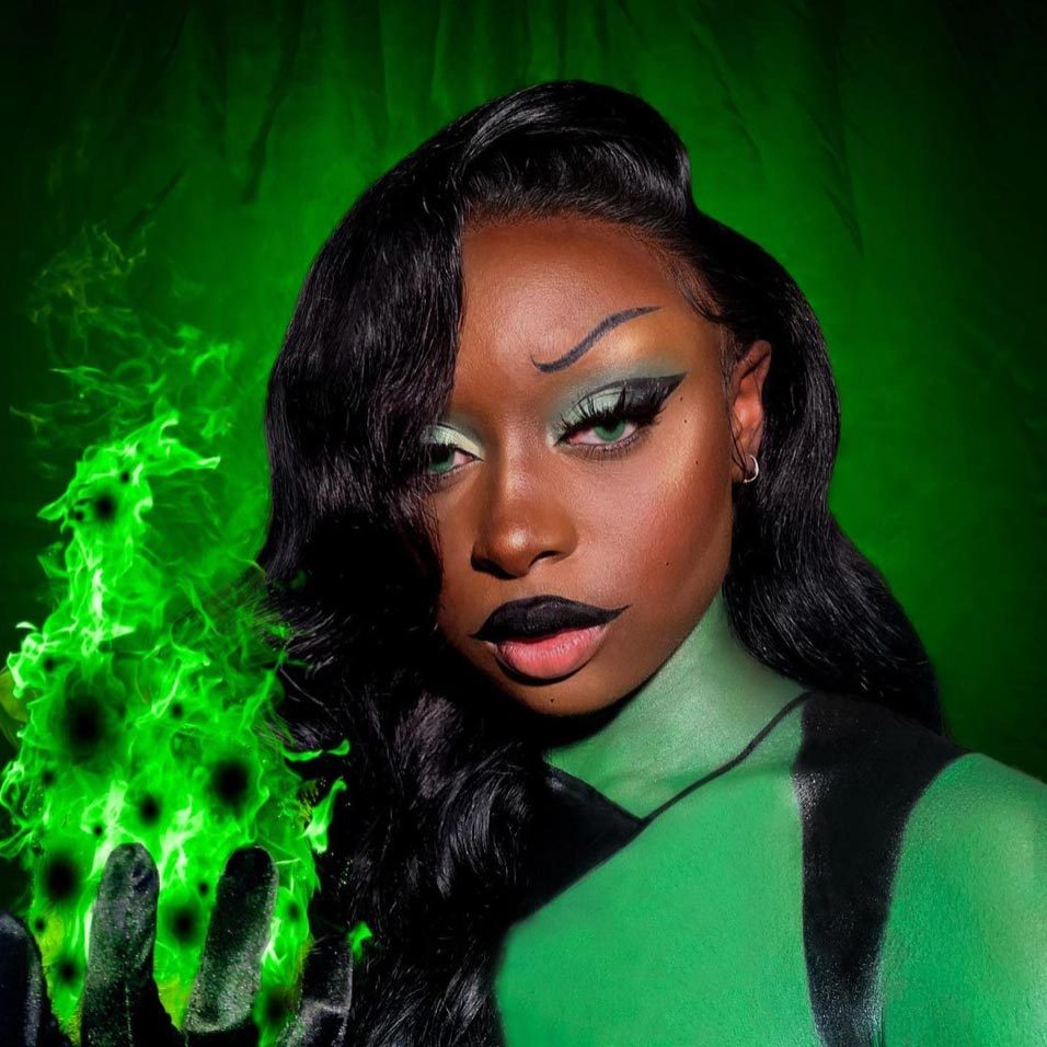 Person holds glowing green flames against a matching green backdrop, with dramatic makeup and dark clothing enhancing the intense visual effect.