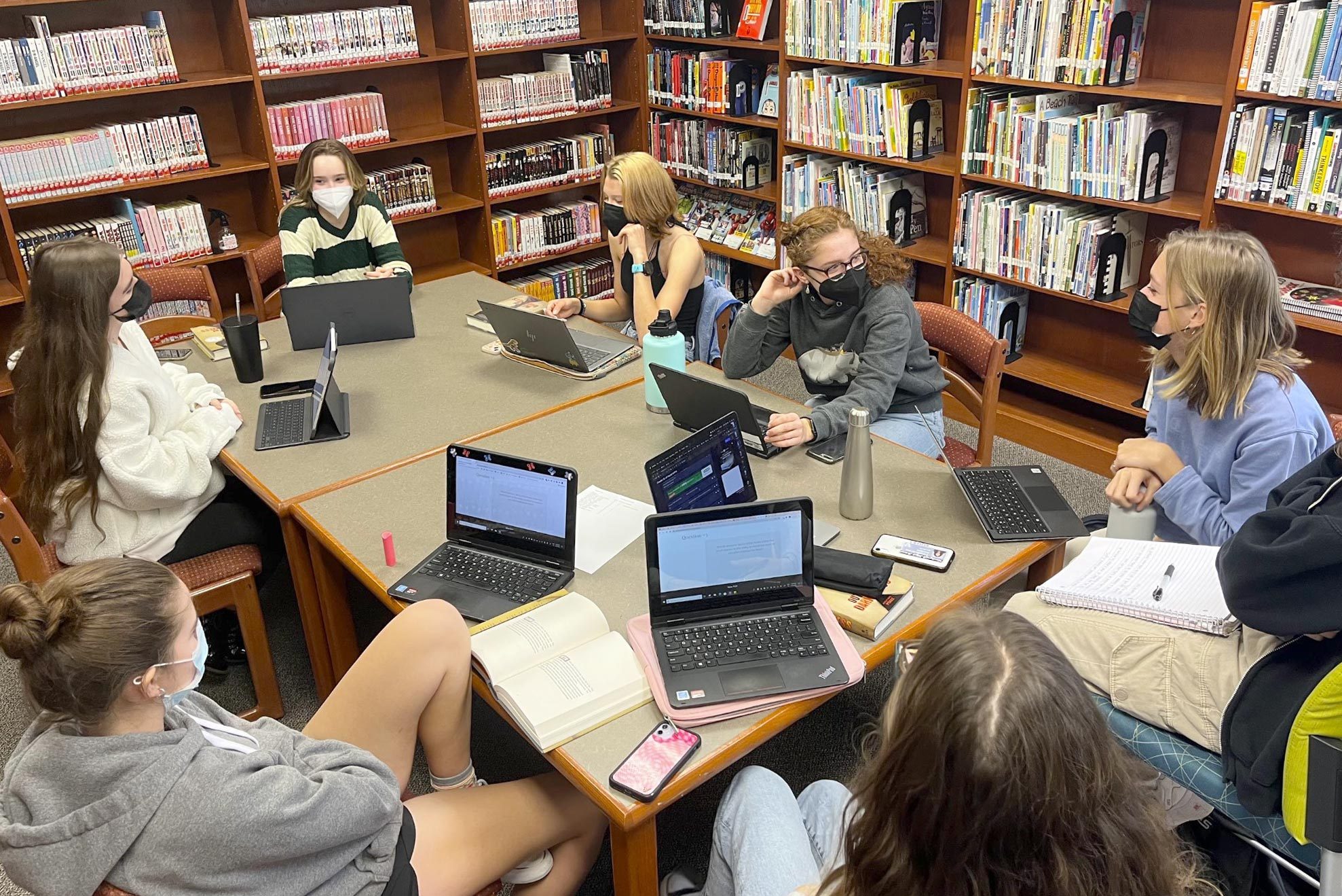 a group of teens at school sitting around a table at the library with laptops for book club
