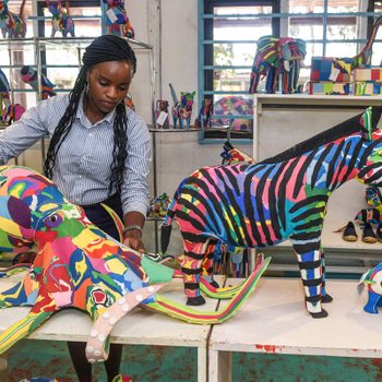 Colorful animal sculptures displayed, with a woman arranging them in a room filled with vibrant, artistic decorations and natural light coming through windows.