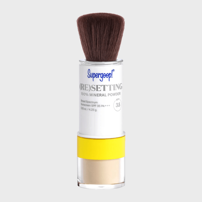 A makeup brush dispenses setting powder from a yellow-labeled container with 