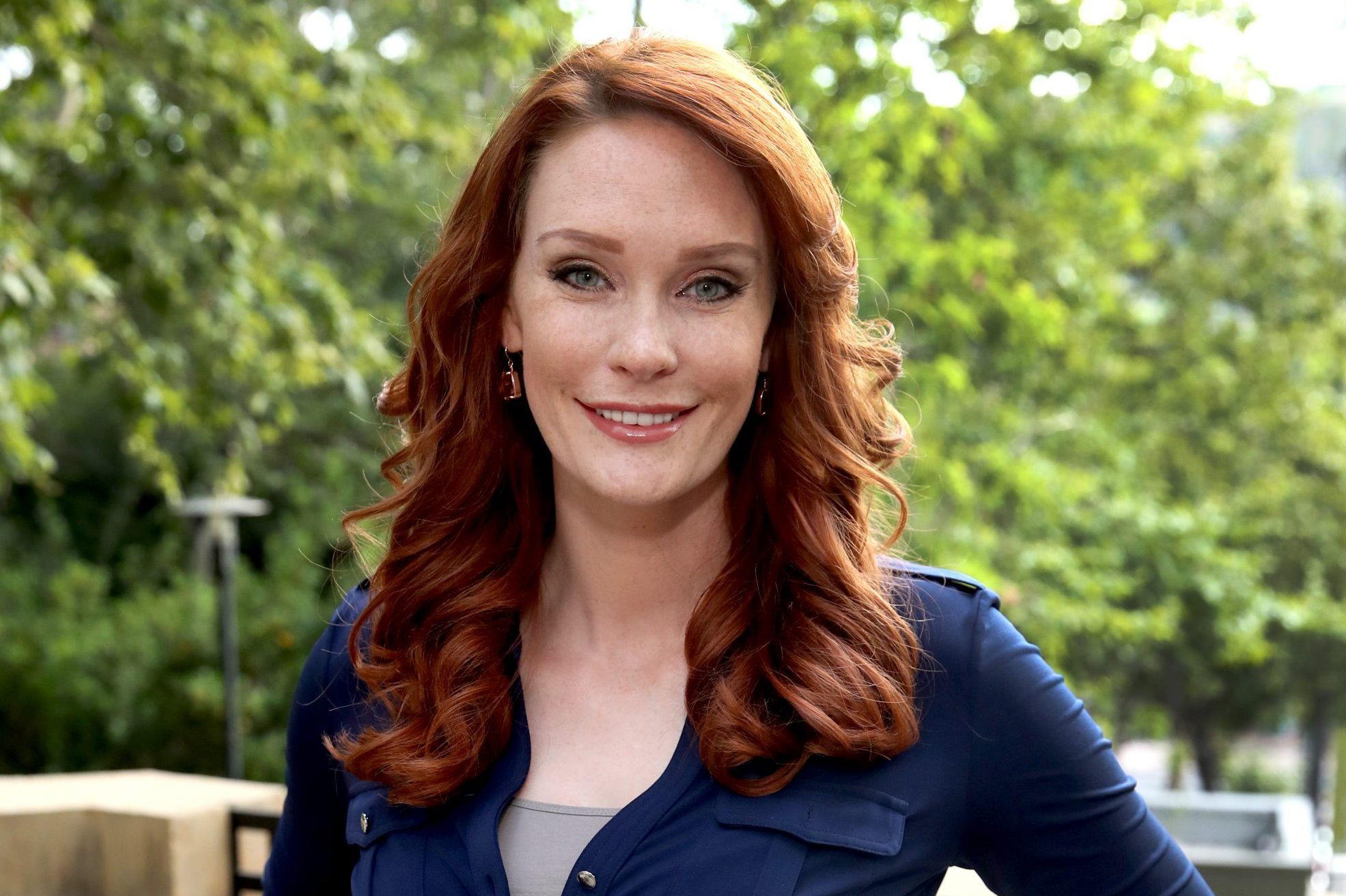 A person with red hair smiling while standing outdoors, surrounded by lush green trees.