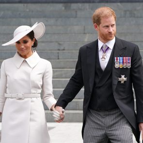Two people hold hands, walking down steps. One wears a white coat and hat, the other a black suit adorned with medals, in an outdoor setting.