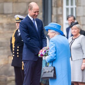 An elderly woman in blue gives flowers to a tall man in a suit, with officials observing in an outdoor, formal setting.