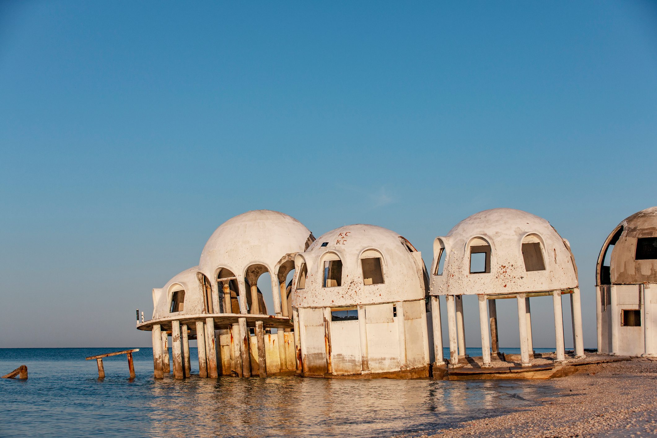 Domed structures stand partially submerged in water, surrounded by a calm, open sea and clear blue sky.