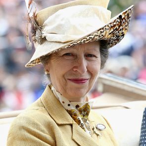 Individual in a feathered hat smiles, wearing a patterned shirt and brooch, outdoors with a blurred crowd in the background.