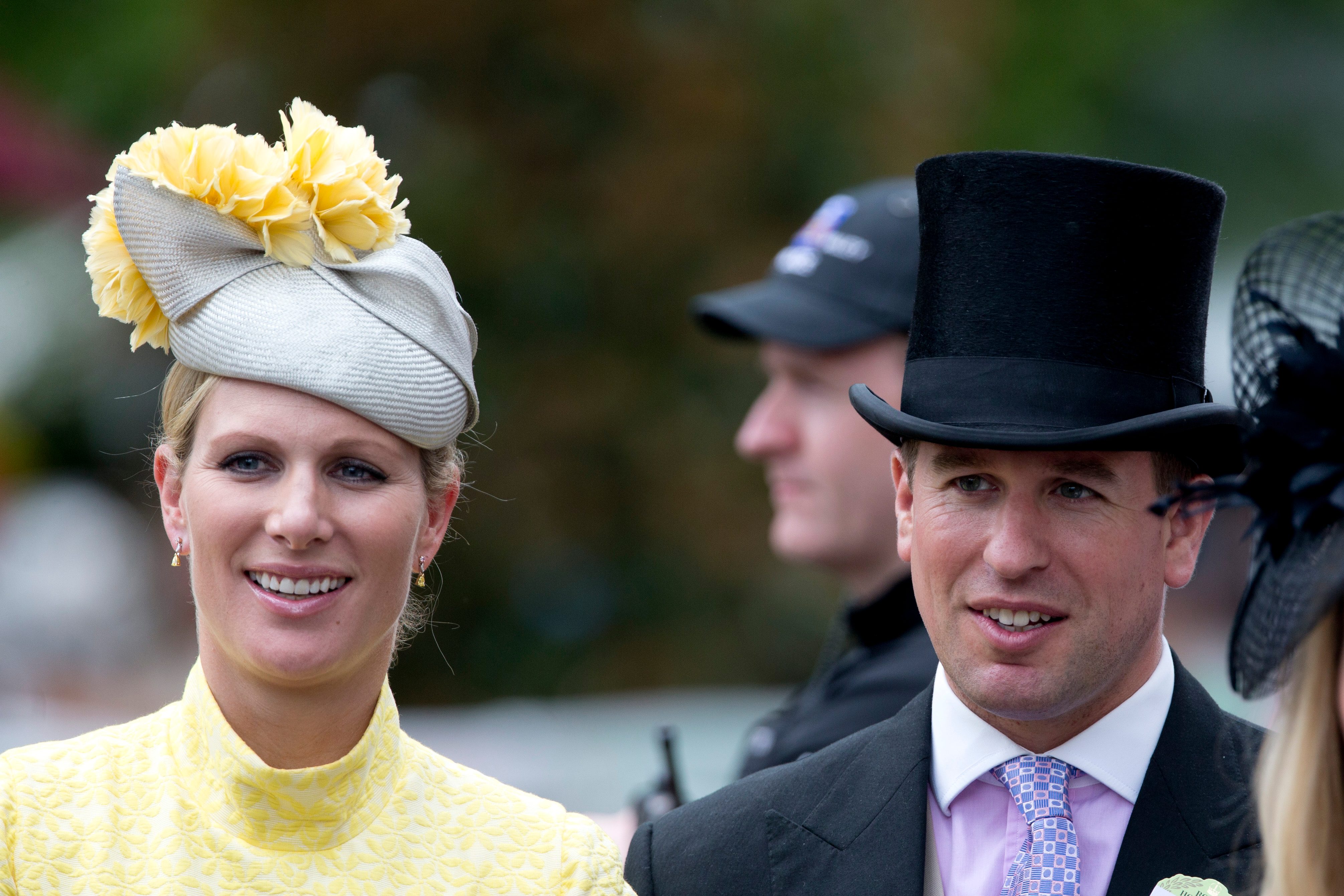 Two people smile, dressed in formal attire with hats, in an outdoor setting with blurred background and other individuals partially visible.