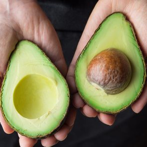 Two avocado halves held in hands, one with a seed, the other without, against a dark background.