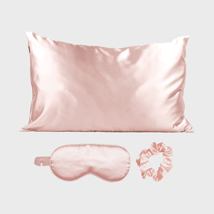 Silk pillowcase rests above matching eye mask and scrunchie on a gray background.