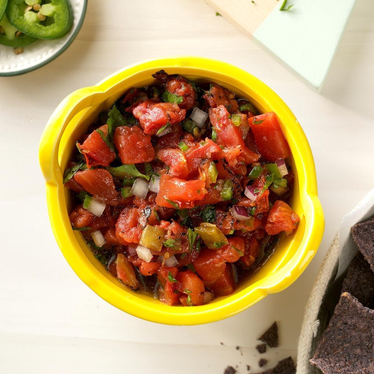 Salsa sits in a vibrant yellow bowl, surrounded by sliced jalapeÃ±os on a plate and a basket of blue corn tortilla chips.