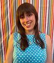 A person smiles, wearing a blue polka dot dress, standing against a colorful, striped curtain background.
