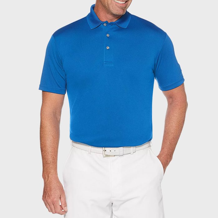 Man wearing a blue polo shirt and white pants stands smiling, hands in pockets, against a plain gray background.