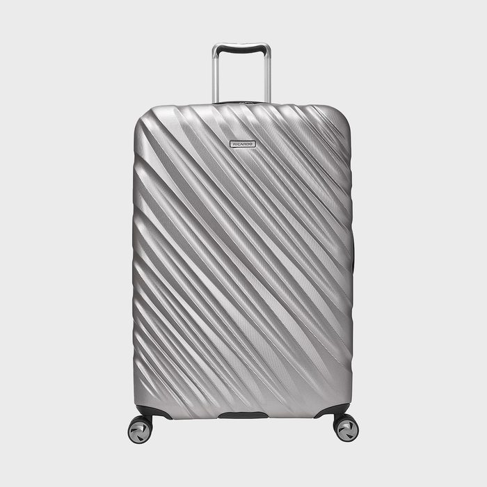 Silver suitcase stands upright; textured with diagonal ridges, featuring a handle and wheels, against a plain white background. Label reads: 
