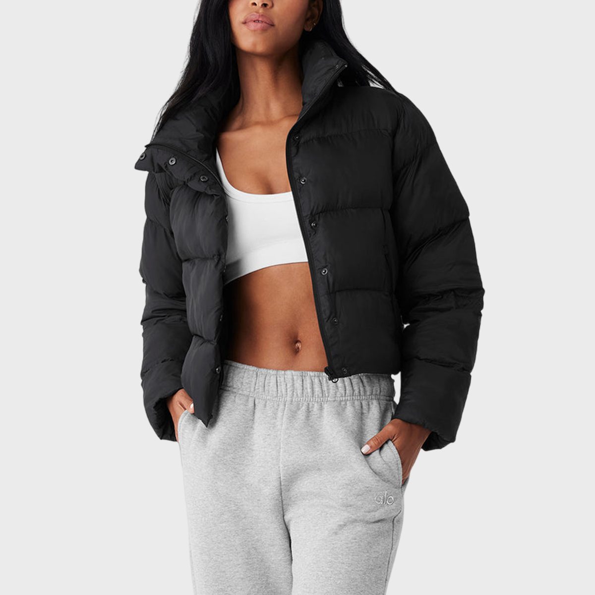 A person wears a black puffer jacket and gray sweatpants, with hands in pockets against a plain background.
