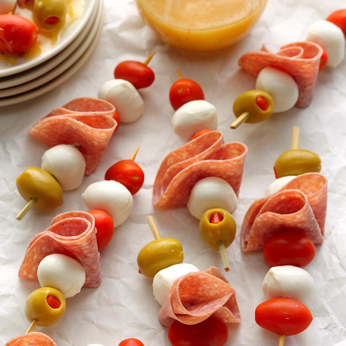 Skewers display stacked salami, mozzarella, tomatoes, and olives on crinkled parchment; a sauce dish sits nearby, enhancing the appetizer presentation.