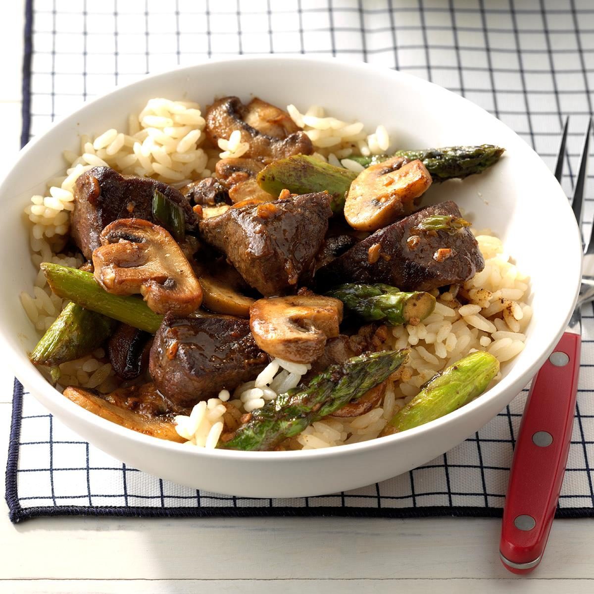 Bowl holds sautÃ©ed beef, mushrooms, asparagus over white rice, placed on a checkered cloth with a red-handled fork nearby.