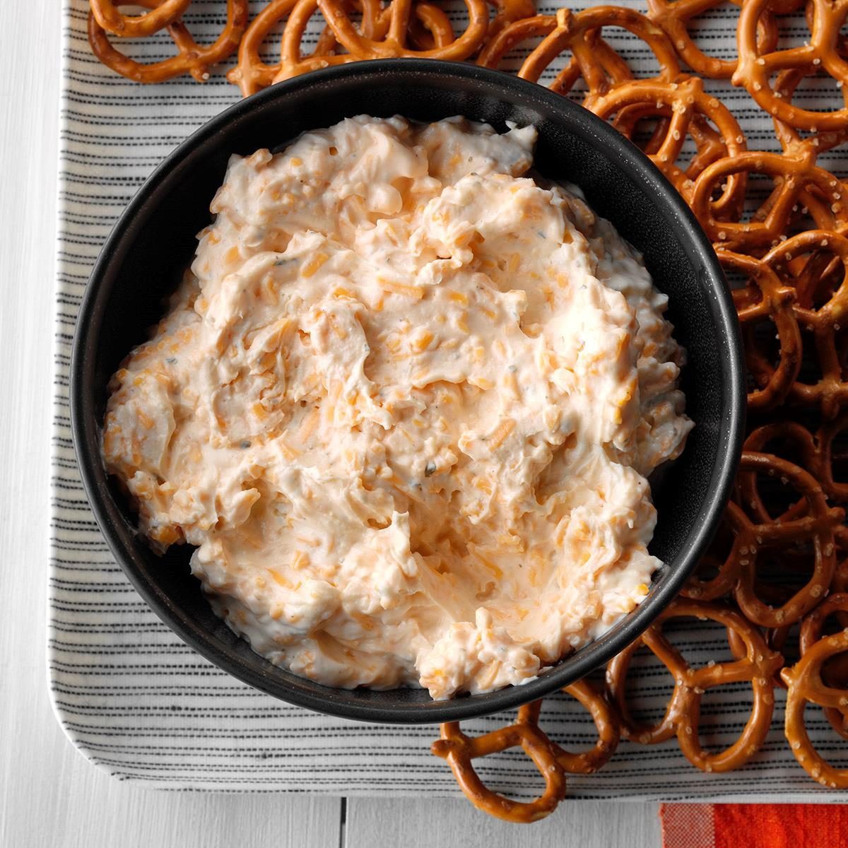 A bowl holds creamy dip, surrounded by pretzels on a striped, rectangular tray.