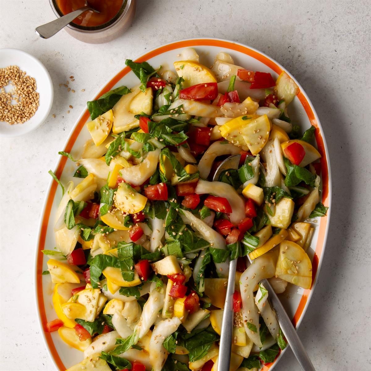 Salad with vibrant vegetables sits garnished on a white plate with orange trim; a jar and bowl of sesame seeds nearby on a gray surface.