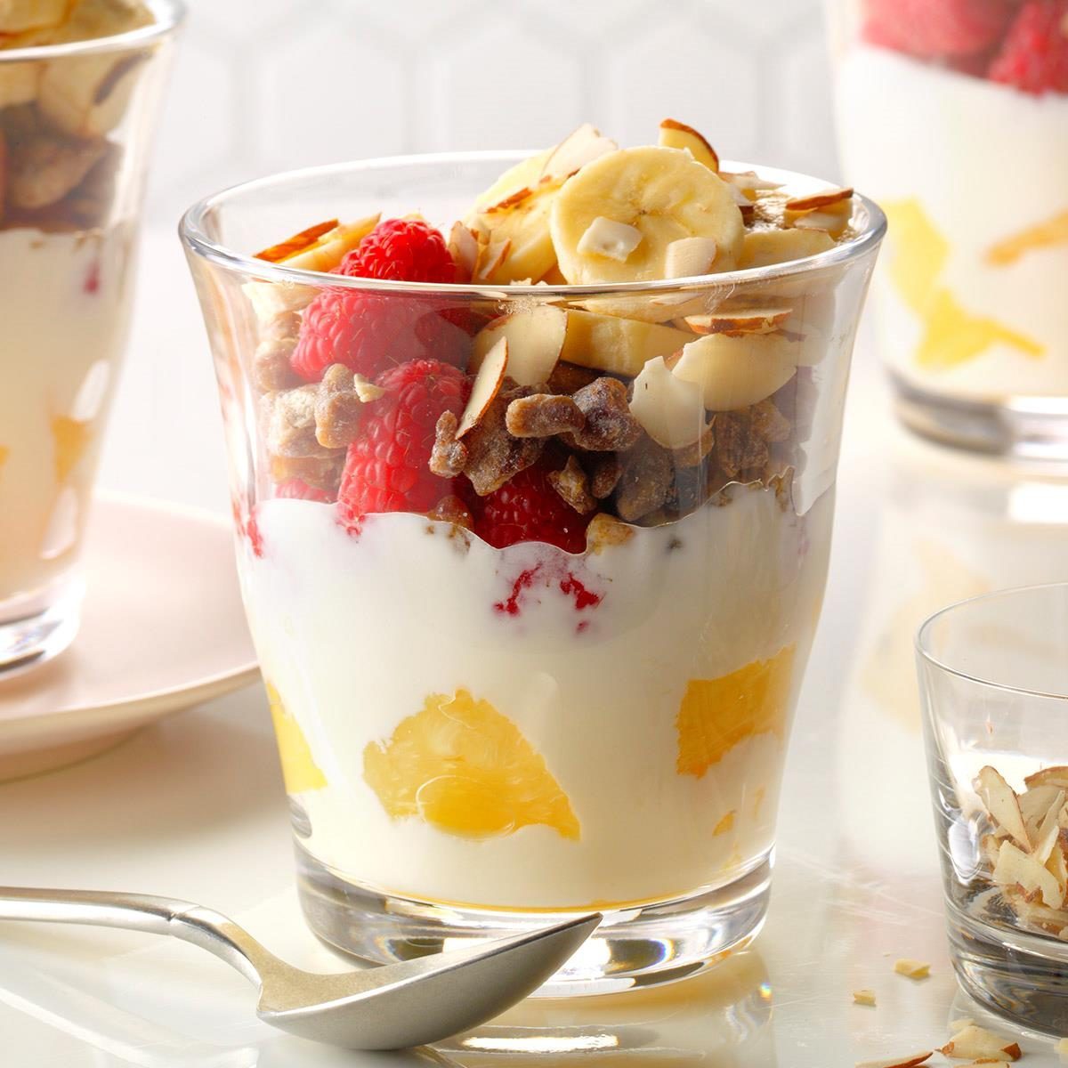 A glass cup holds layered yogurt, raspberries, banana slices, nuts, and pineapple chunks on a light, reflective surface with a spoon nearby.