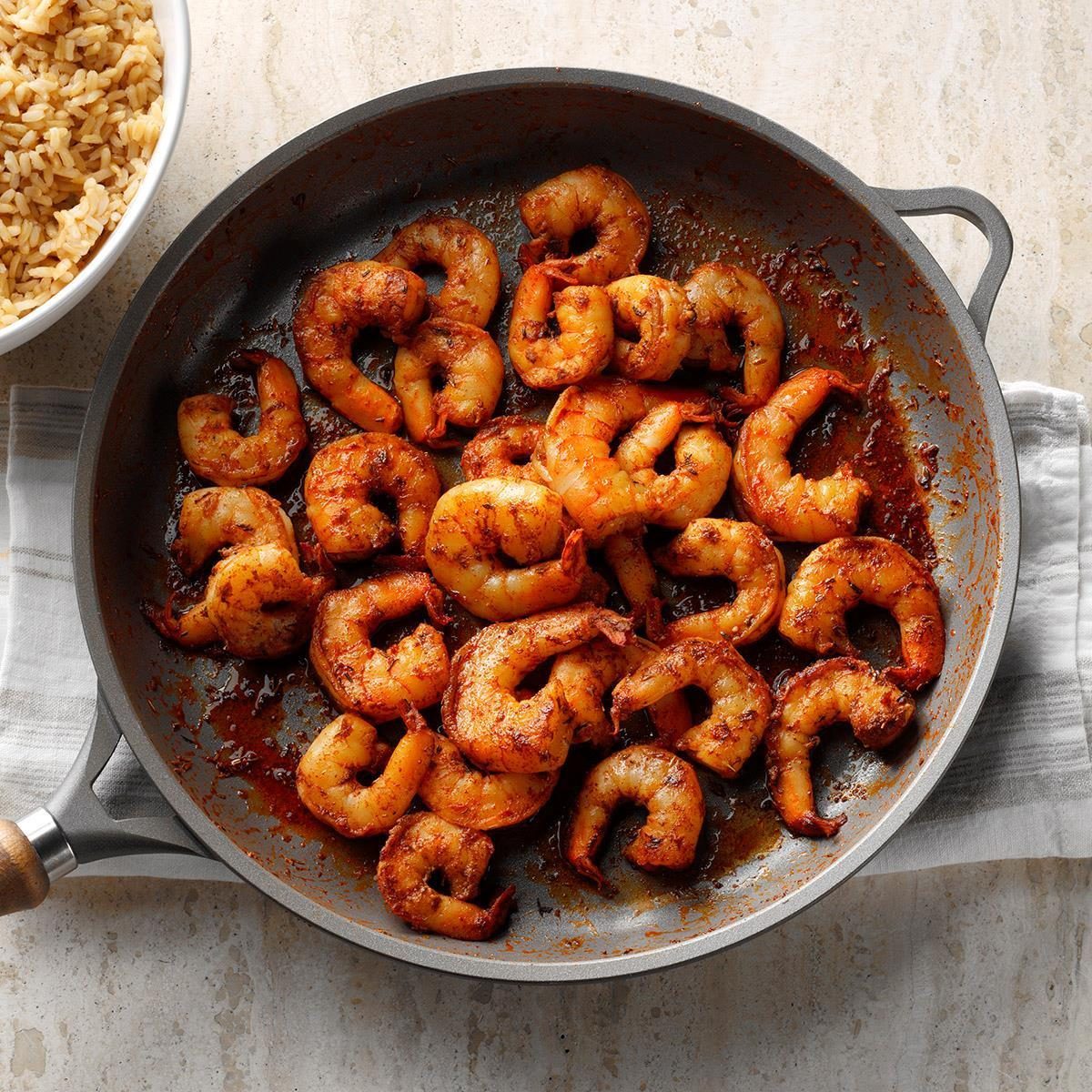 Shrimp are sizzling in a pan on a striped towel, with a bowl of rice nearby on a light countertop.
