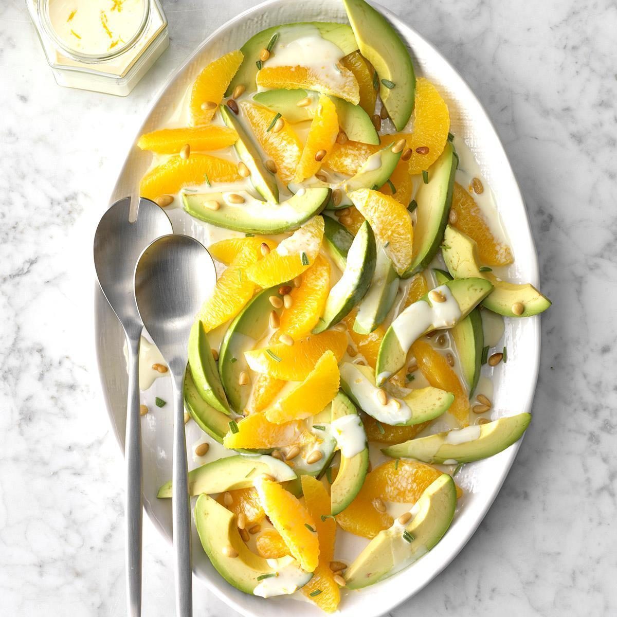 Salad with avocado and orange slices arranged on a white plate, drizzled with dressing and topped with seeds; two serving spoons lie beside it.