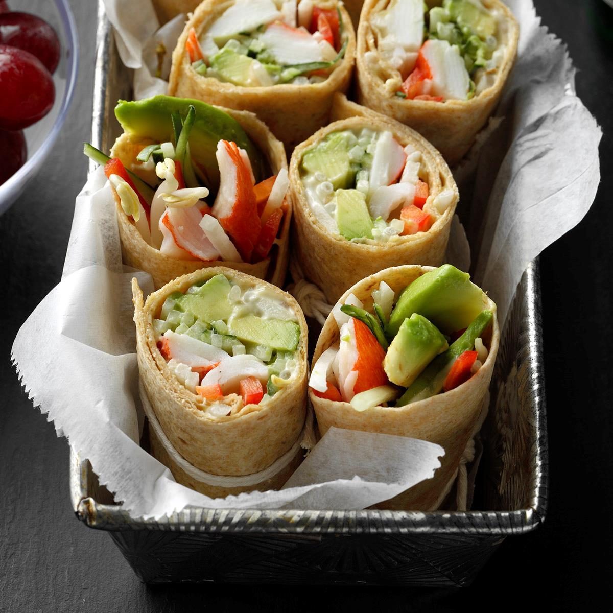 Wraps filled with vegetables and crab sticks are neatly arranged in a small tray lined with parchment paper, next to a bowl of grapes.