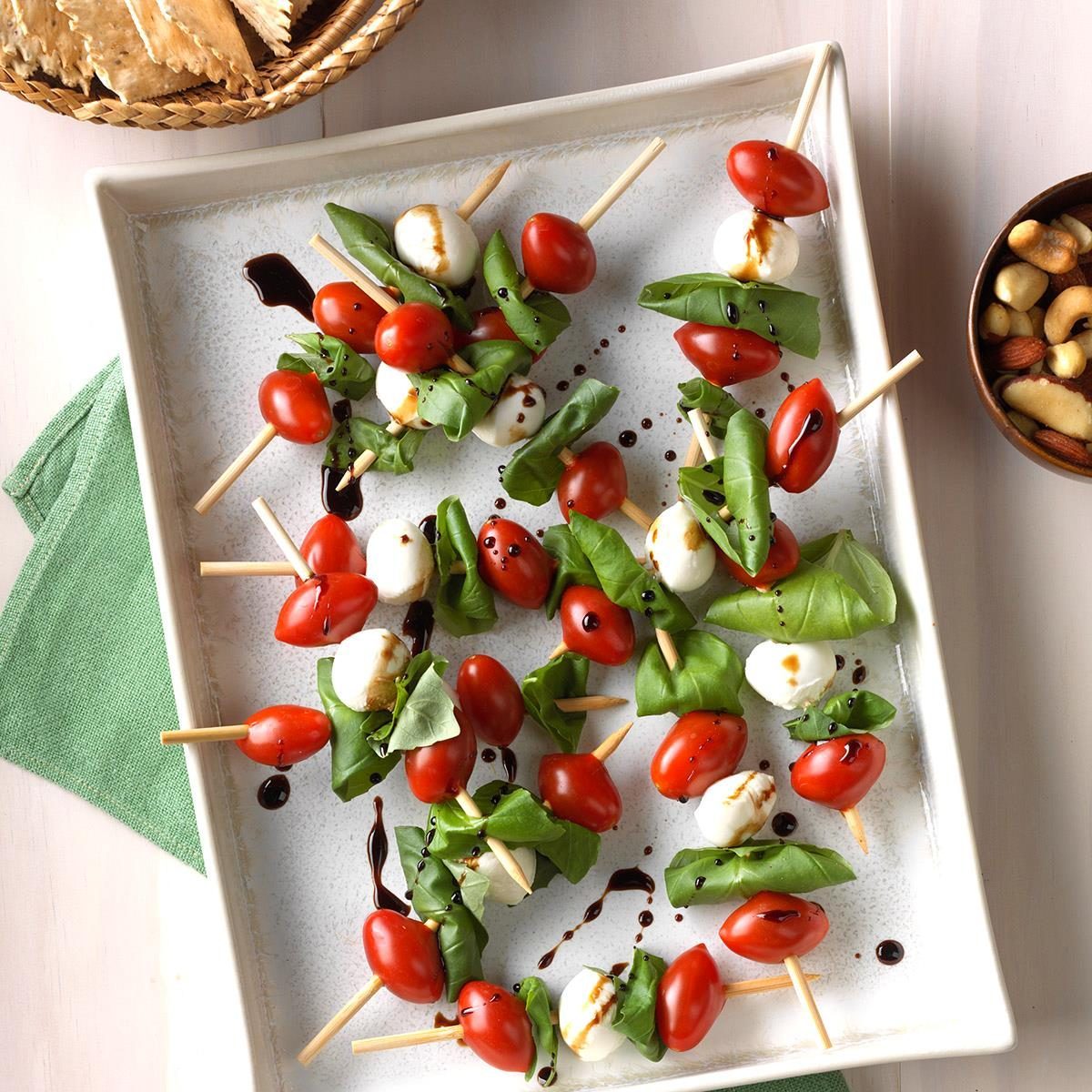 Skewers hold cherry tomatoes, mozzarella, and basil drizzled with balsamic glaze on a white dish; nuts and crackers are in bowls nearby.