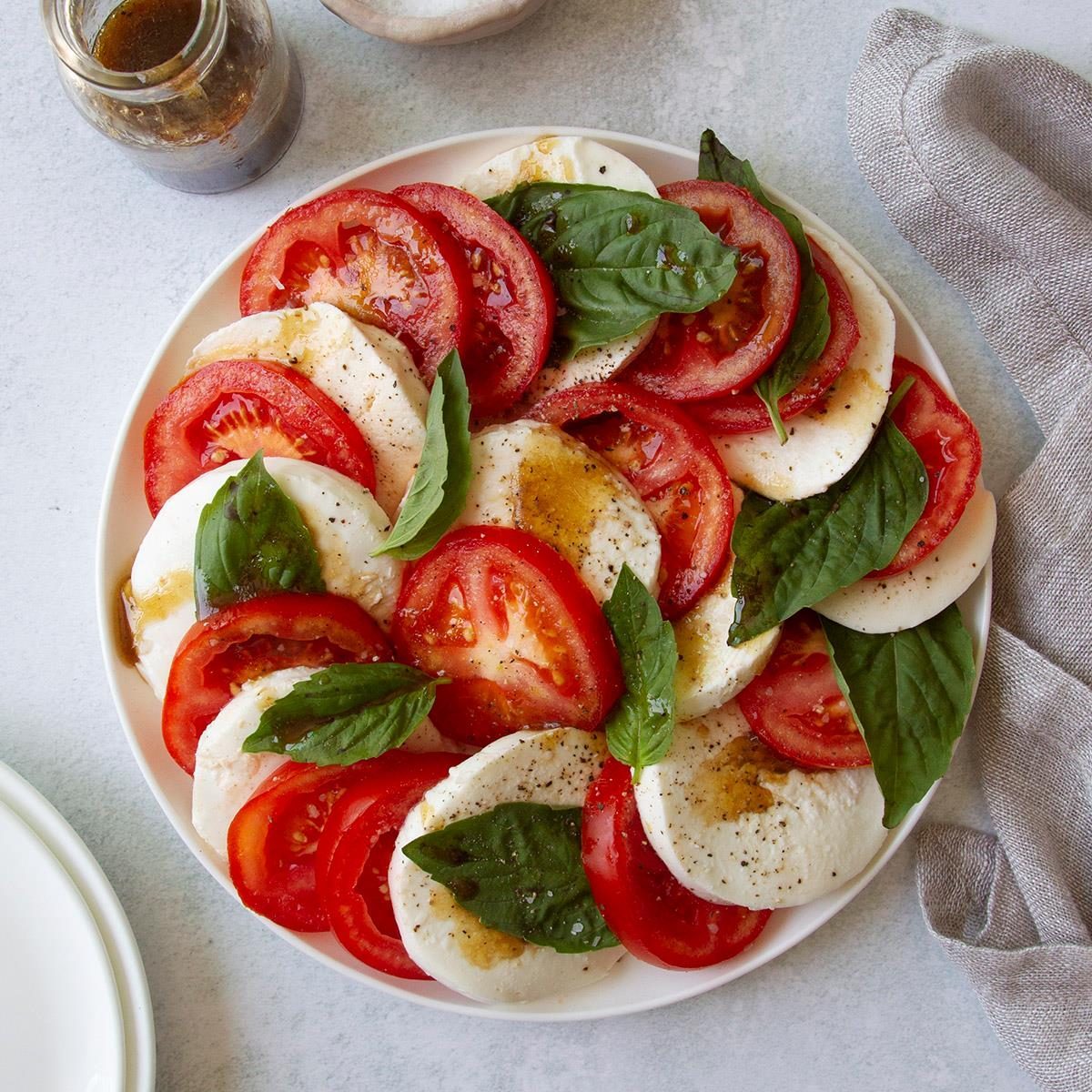 Caprese salad with sliced tomatoes, mozzarella, and basil, drizzled with balsamic dressing, on a white plate with a gray cloth nearby.