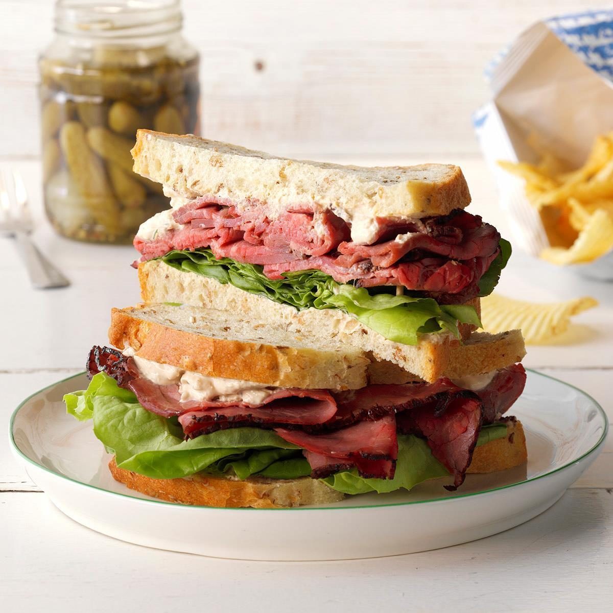 Sandwich stacked high with layers of roast beef and lettuce, sits on a white plate; pickles and chips in the background.