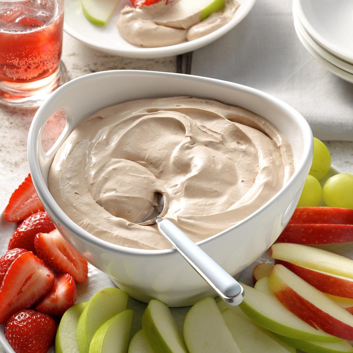 A bowl holds creamy dip with a spoon. Surrounding it are fresh sliced strawberries, apples, and grapes on a white platter.