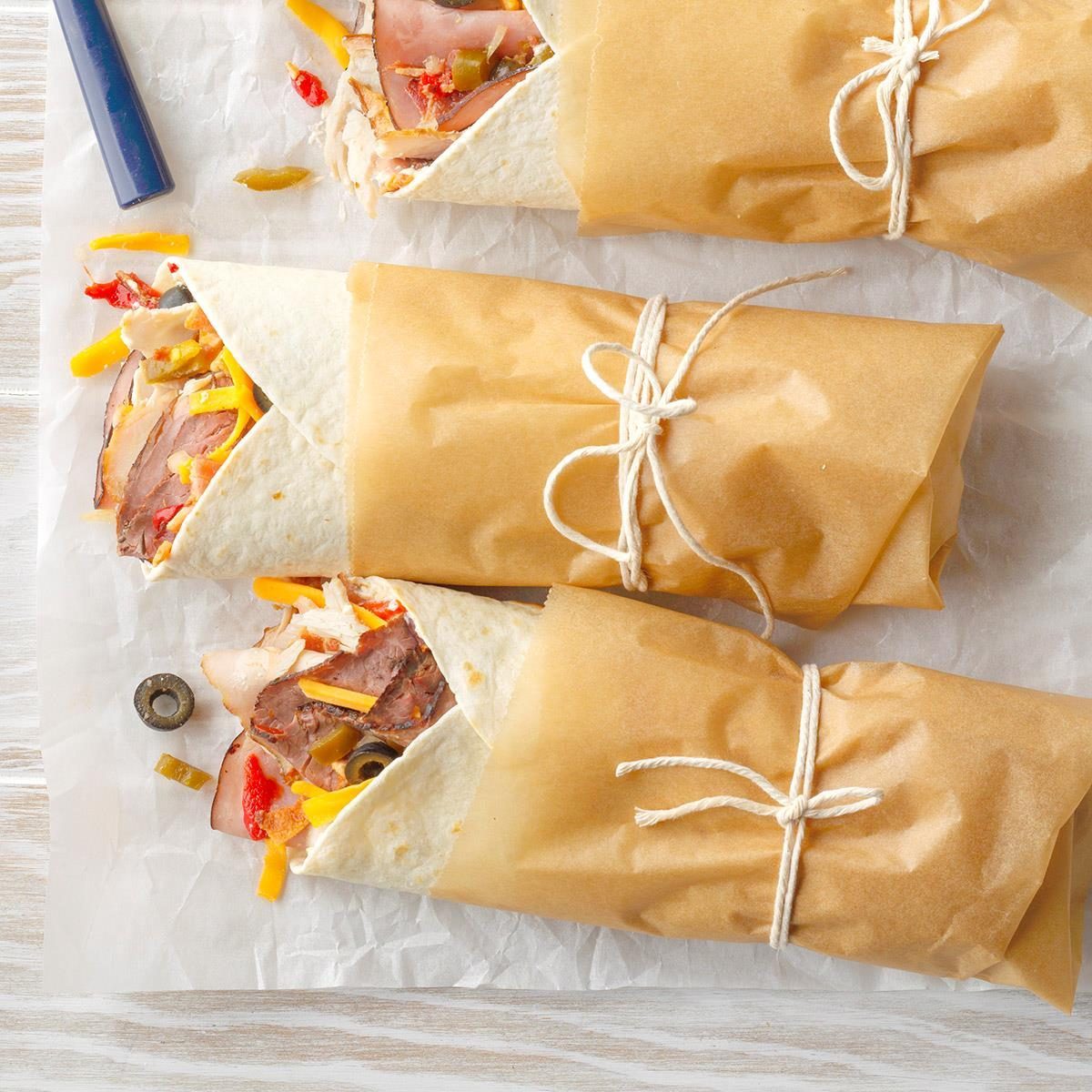 Three wrapped sandwiches tied with string rest on parchment paper, filled with meats and vegetables, near a pen and an olive.