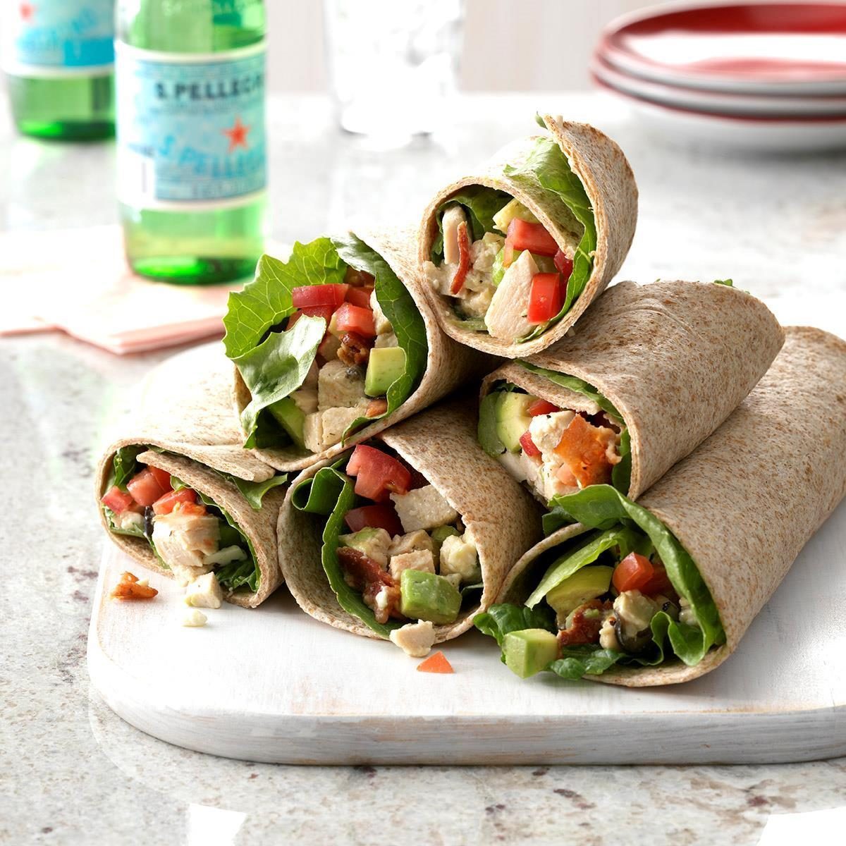 Wraps filled with lettuce, tomato, avocado, and chicken, neatly arranged on a cutting board, with bottled drinks and plates visible in the background.