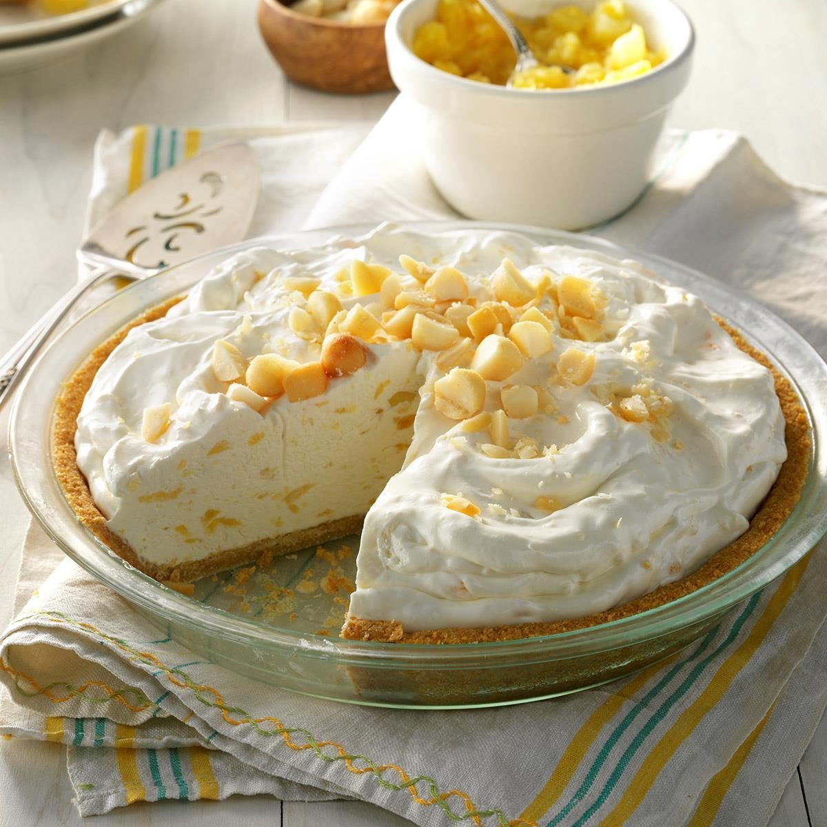 Pie sits on a table surrounded by a striped cloth, with a slice removed. Itâ€™s topped with chopped nuts and adjacent to a white bowl.