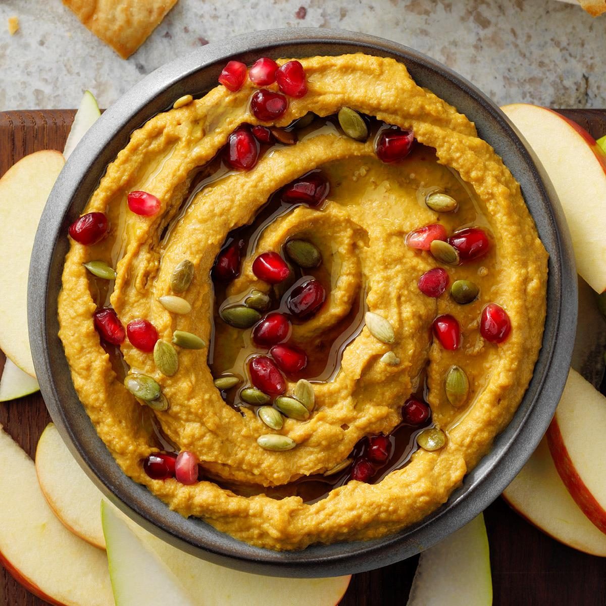 Bowl holding hummus, garnished with pomegranate seeds and pumpkin seeds, surrounded by sliced apples on a textured surface.