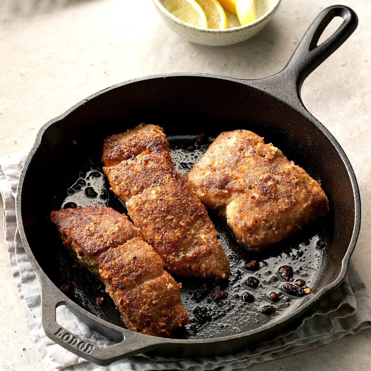 Pan-seared fish fillets sizzle in a cast-iron skillet on a striped cloth with lemon wedges beside it.