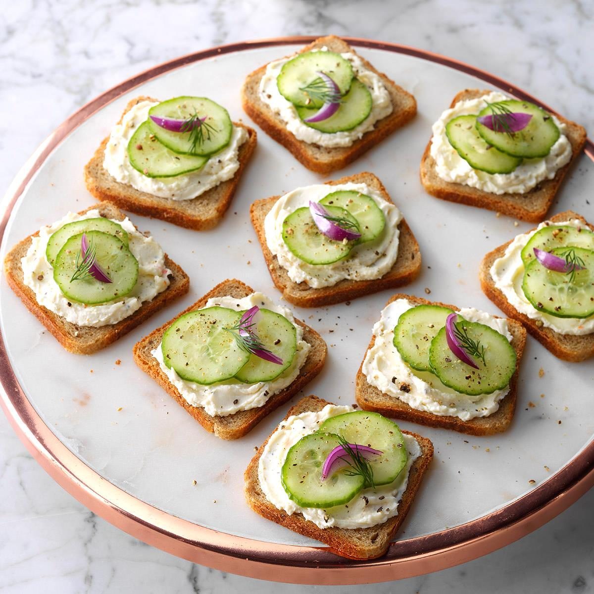 Toast squares topped with cream cheese, cucumber slices, and herbs sit on a round marble platter in a bright kitchen setting.
