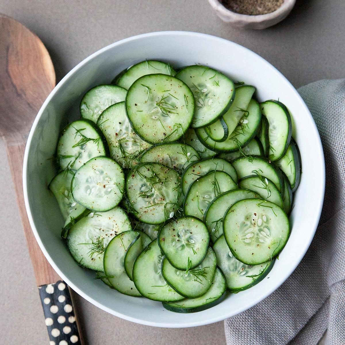 Cucumber slices rest in a white bowl, sprinkled with dill and spices, beside a wooden spoon on a textured fabric surface.